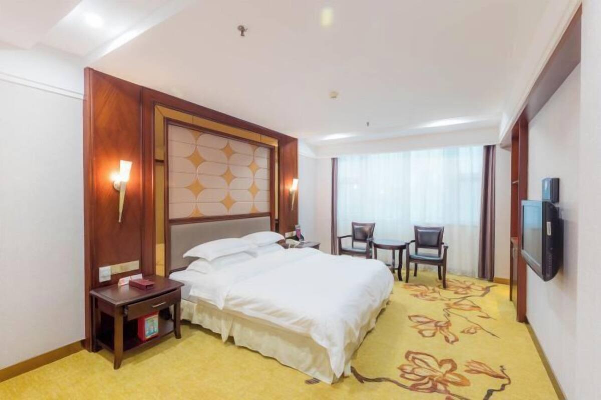 Een hotelkamer met een groot bed, een nachtkastje, een stoel, een televisie en een raam met gordijnen.