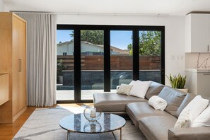 Ein modernes Wohnzimmer mit einem Ecksofa, einem Glastisch und Blick durch große Schiebetüren auf einen Außenbereich.