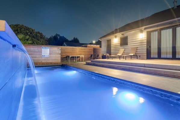 Una piscina con cubierta de madera y sillas de descanso, rodeada por un edificio con grandes ventanales y un patio cubierto.