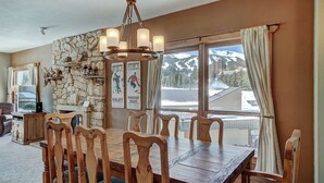Un comedor con mesa y sillas de madera, pared de piedra, candelabro y vista a montañas a través de amplios ventanales.