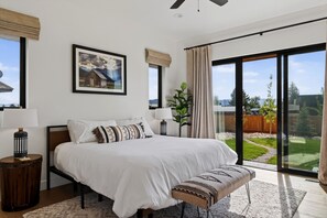 A bedroom with a large bed, a bench, a nightstand, a lamp, a framed picture, and a view of a backyard through a sliding glass door. - New Modern 5BR with AC - Walk to Targhee Shuttle (Driggs)