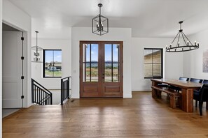 A spacious room with a wooden floor, a dining table with chairs, a staircase with black railings, and a large wooden door with a view. - New Modern 5BR with AC - Walk to Targhee Shuttle (Driggs)