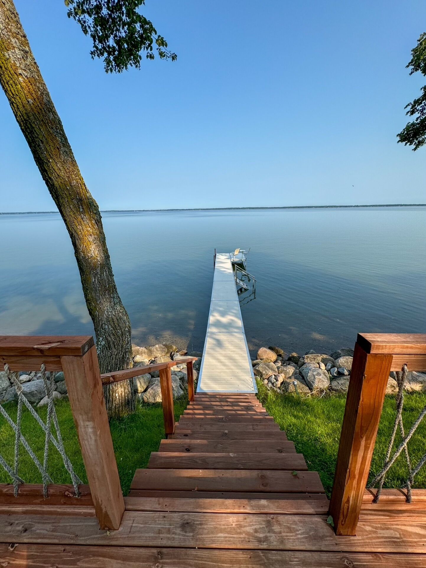 A wooden staircase leading to a dock over calm water, surrounded by greenery and a clear sky.