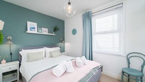 Un dormitorio ordenado con cama, mesitas de noche, una silla y un ventanal con cortinas.