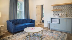A living room with a blue sofa, a small round coffee table, and a patterned rug. There is a kitchen area with a microwave and a shelf above the counter.