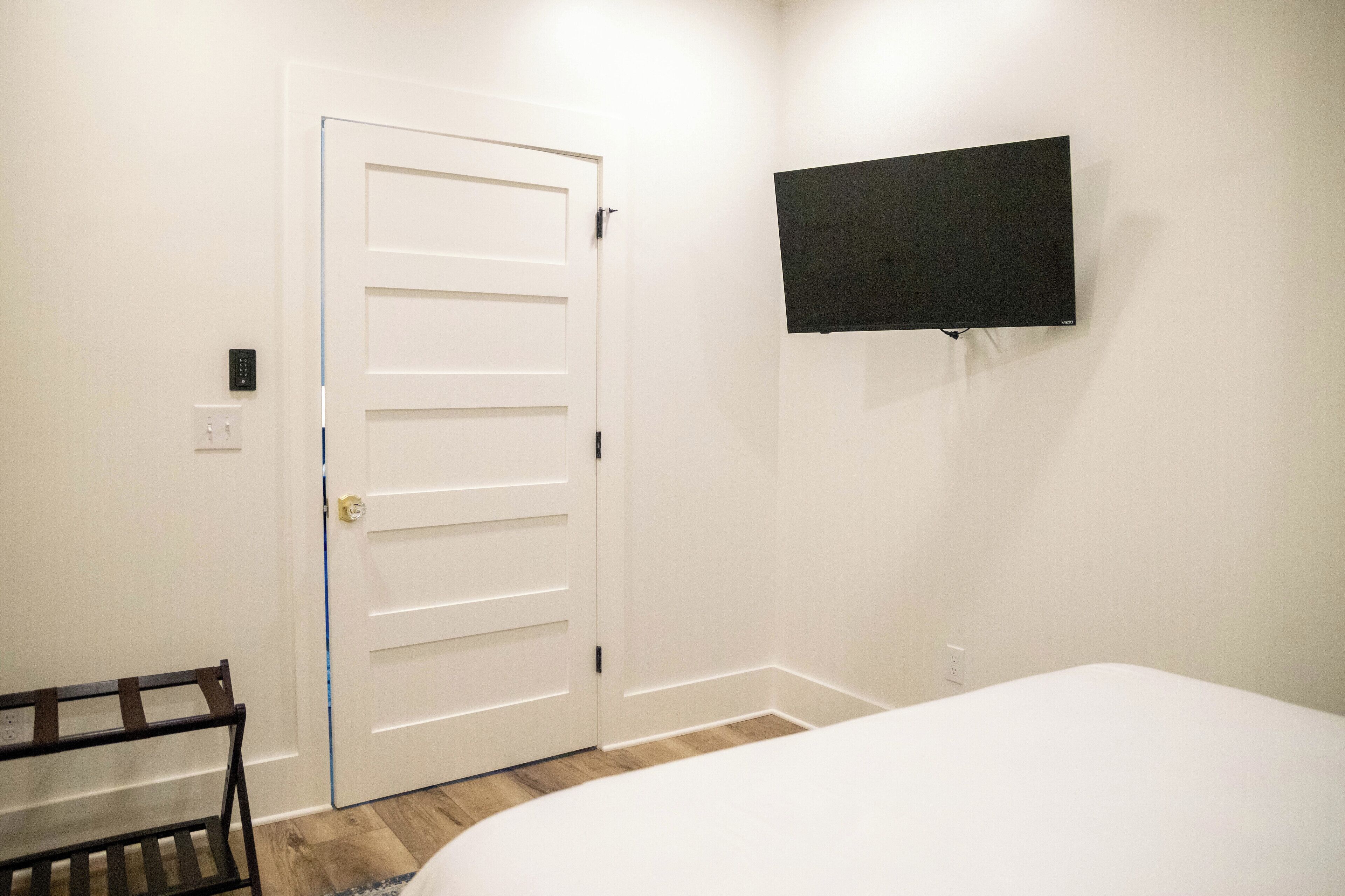 Un dormitorio con una puerta blanca, televisor montado, banca de madera y una cama.