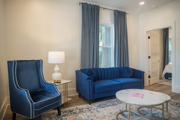 Una habitación con un sillón azul, un sofá azul, una mesita con una lámpara y una alfombra estampada.