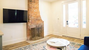 A living room with a brick fireplace, a flat-screen TV mounted on the wall, a round coffee table, and a patterned rug.