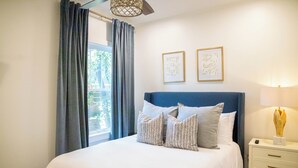 A neatly made bed with a blue headboard, a bedside table with a lamp, and two framed artworks on the wall.