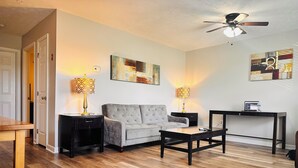 Un salon avec un canapé gris, une table basse, deux tables d’appoint avec des lampes, un ventilateur de plafond et des œuvres d’art accrochées aux murs.