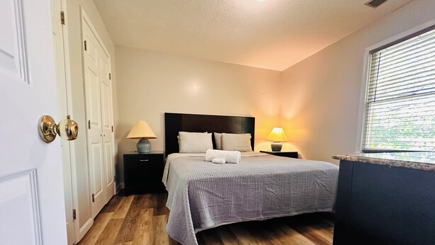 Une chambre à coucher avec un lit, deux tables de chevet avec des lampes, un plancher en bois et une fenêtre avec des stores.