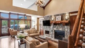 A spacious living room with a stone fireplace, comfortable seating, and a large window overlooking a scenic view. - Ski In, Ski Out 3 Bedroom Condo at the Eagle Bahn Gondola With Slopeside Pool (Vail)
