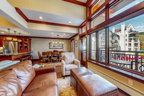 A spacious living room with leather sofas, a dining area, and large windows offering a view of a cityscape. - Ski In, Ski Out 3 Bedroom Condo at the Eagle Bahn Gondola With Slopeside Pool (Vail)