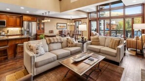 A spacious living area with a large window, a kitchen with wooden cabinets, and a seating area with a coffee table. - Ski In, Ski Out 3 Bedroom Condo at the Eagle Bahn Gondola With Slopeside Pool (Vail)