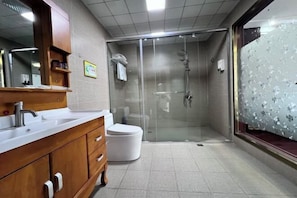 A bathroom with a wooden vanity, a white toilet, a glass-enclosed shower, and a frosted glass window. - Golden Times Holiday Hotel (Huoshan)