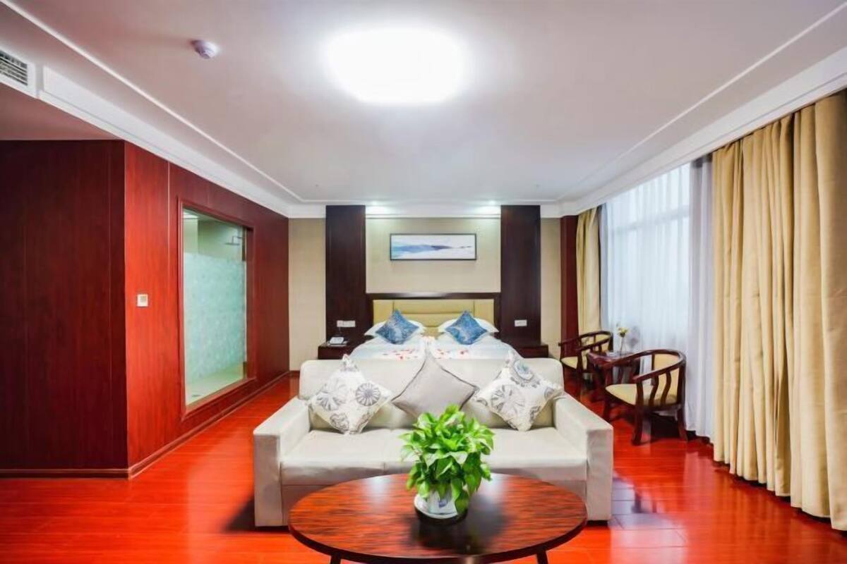 Une chambre d’hôtel avec un canapé, une table à manger, une plante et une grande fenêtre avec des rideaux.