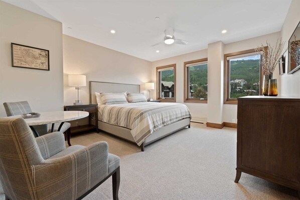 A bedroom with a bed, a desk, two chairs, a dresser, and large windows offering a view of the outdoors. - Beaver Creek Village Ski In, Ski Out 2 Bedroom Residence With Pool (Avon)