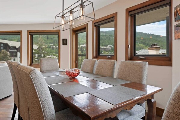A dining room with a wooden table, six chairs, and a bowl of fruit. Large windows offer a view of a house and trees. - Beaver Creek Village Ski In, Ski Out 2 Bedroom Residence With Pool (Avon)