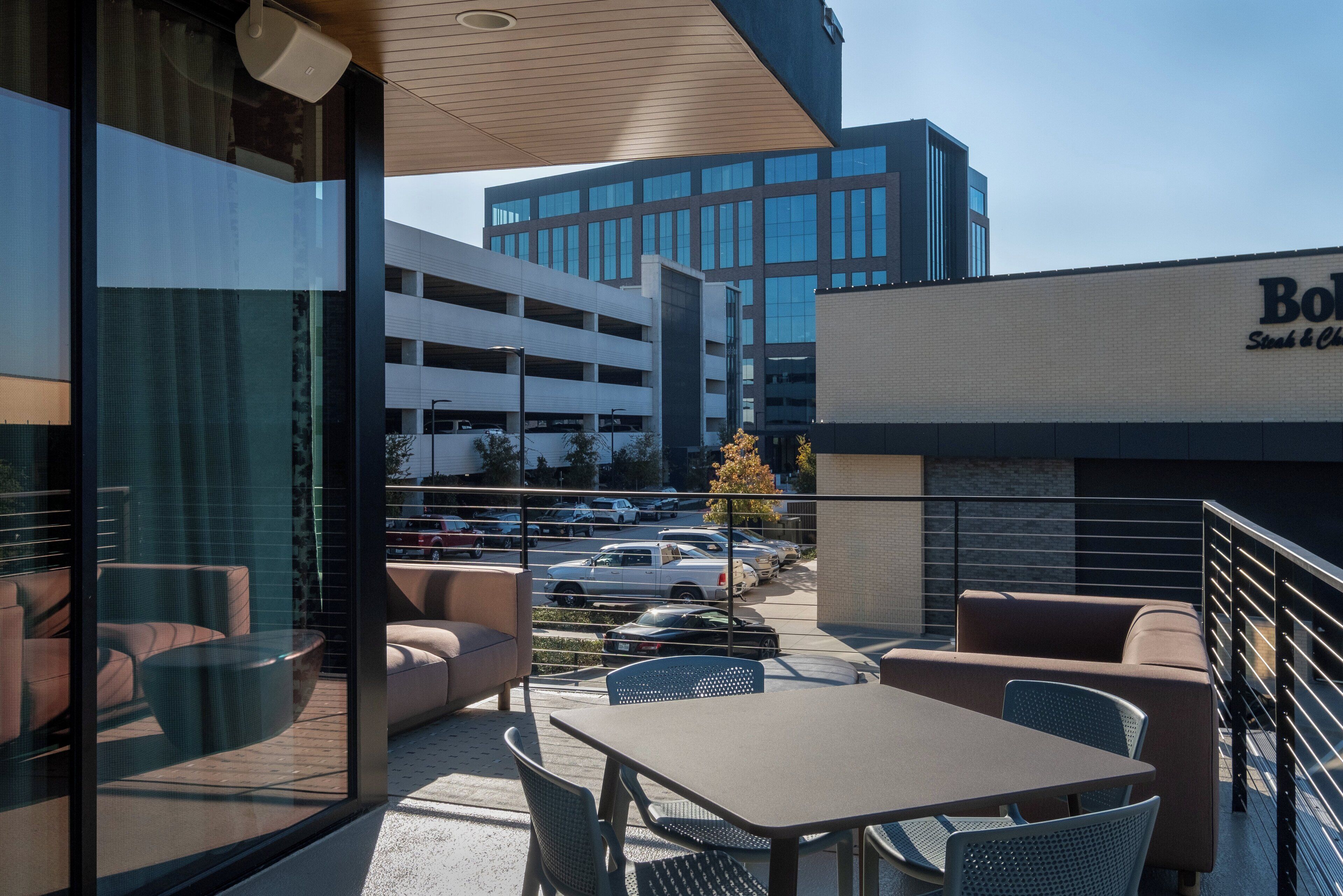 An outdoor seating area with tables and chairs, a glass wall, and a view of a parking garage and a building with a sign.