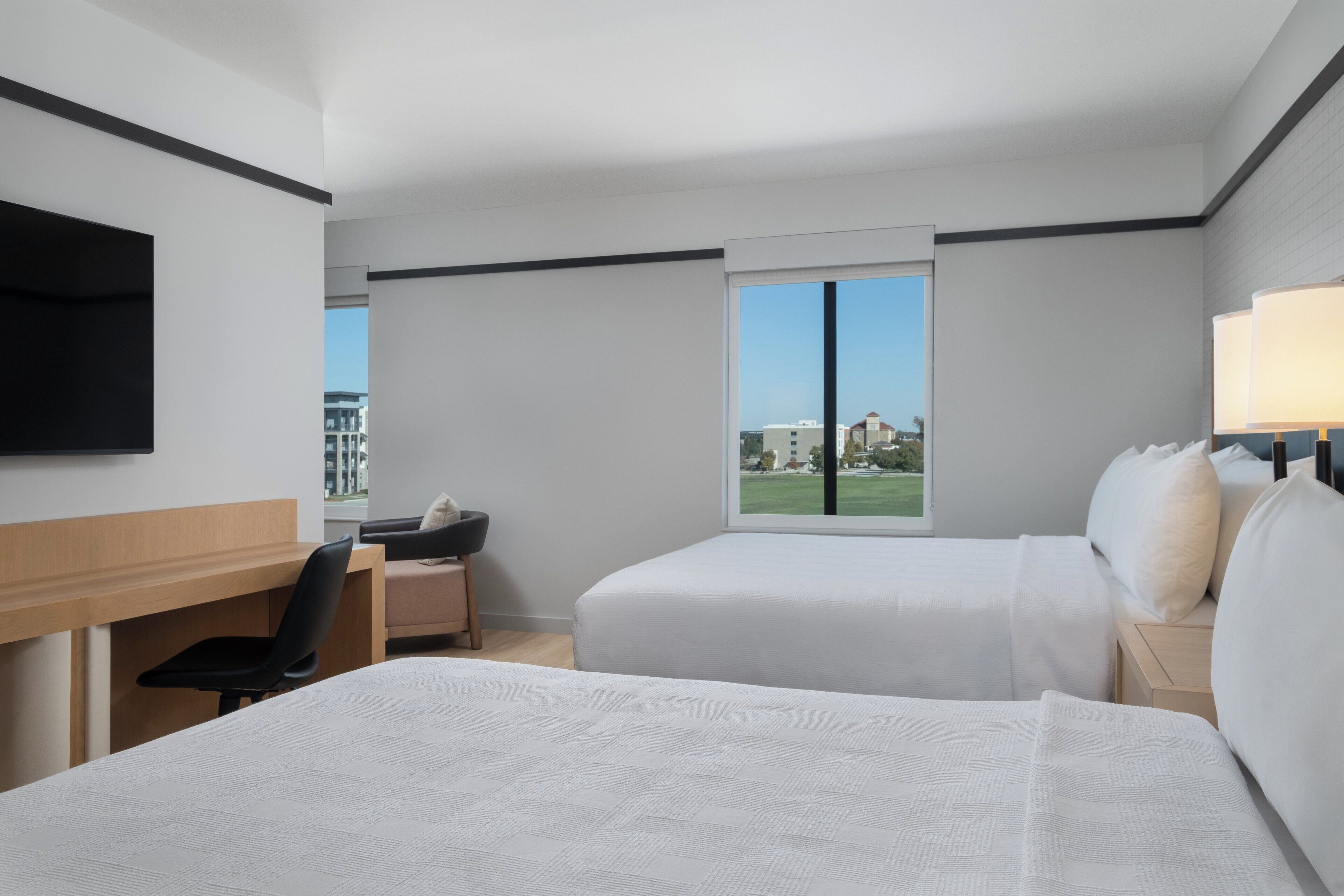 Ein Hotelzimmer mit zwei Betten, einem Schreibtisch, einem Sessel und einem Fernseher. Es gibt ein Fenster mit Blick auf Gebäude und eine Rasenfläche.