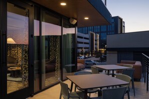 An outdoor seating area with tables and chairs, a glass partition, and a view of a parking garage. - Denizen Hotel (McKinney)