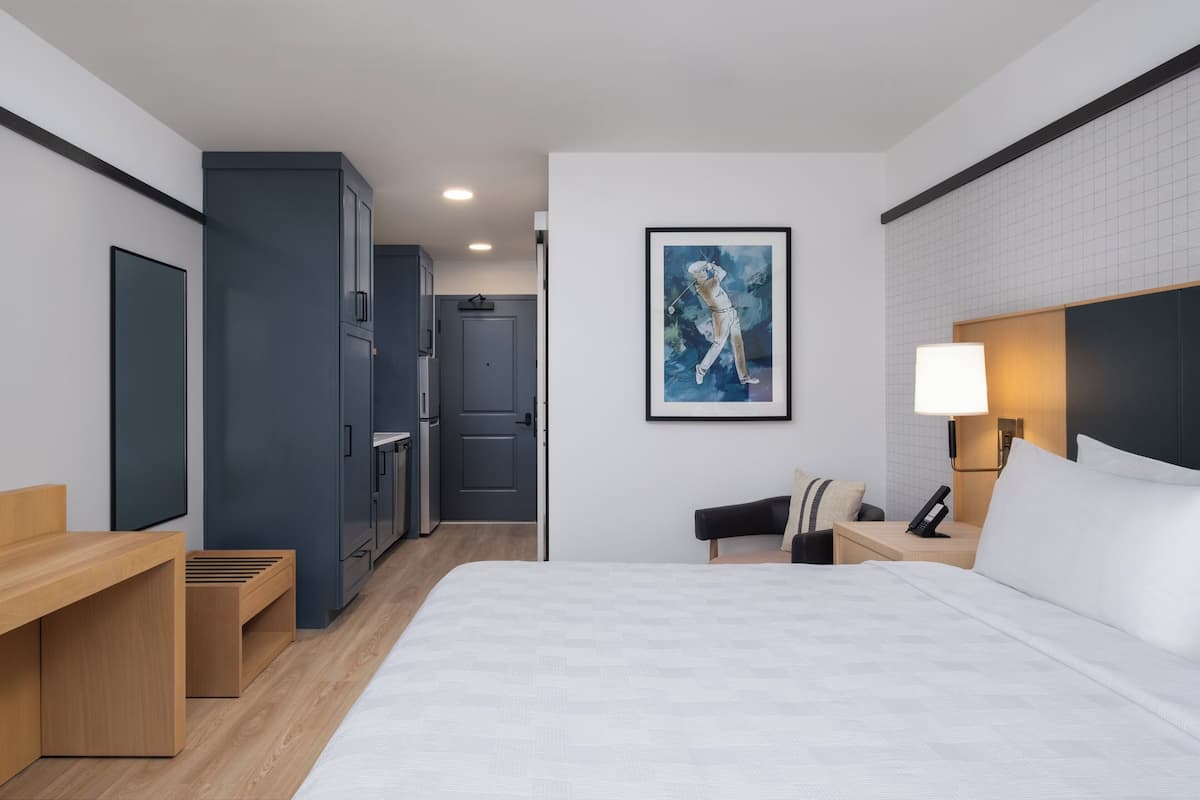A modern hotel room with a large bed, a desk, a chair, a nightstand with a lamp, and a painting on the wall.