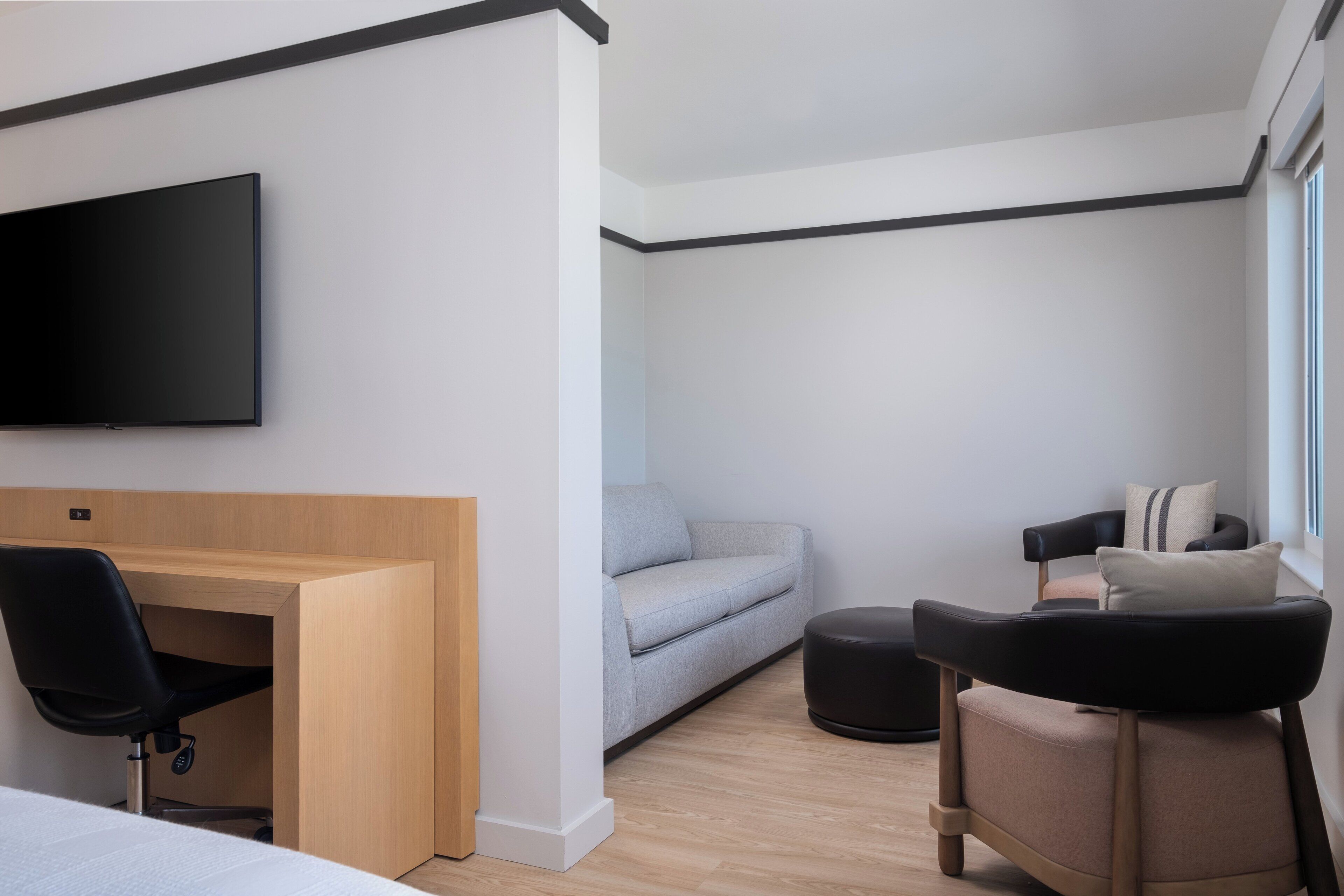 Ein modernes Hotelzimmer mit Flachbildfernseher an der Wand, einem Holztisch mit Stuhl, einem grauen Sofa, einem schwarzen Hocker und einem schwarzen Sessel mit gestreiftem Kissen.