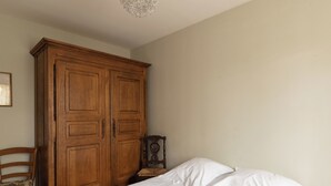 Une chambre avec une grande armoire en bois, un lit avec du linge de lit blanc et une chaise agrémentée d’un coussin à motifs.