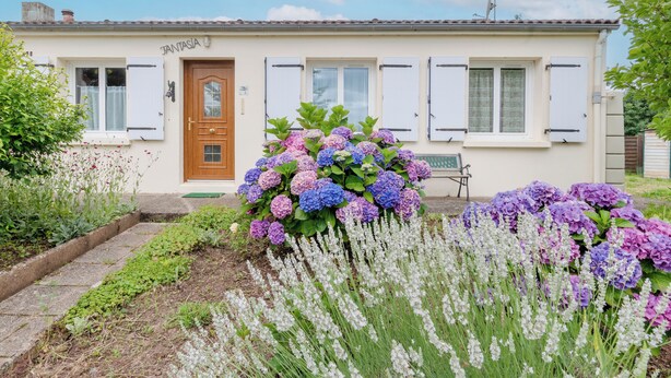 Une maison à deux étages, dotée d’une porte et de volets en bois, entourée d’un jardin où poussent des hortensias et des fleurs blanches.