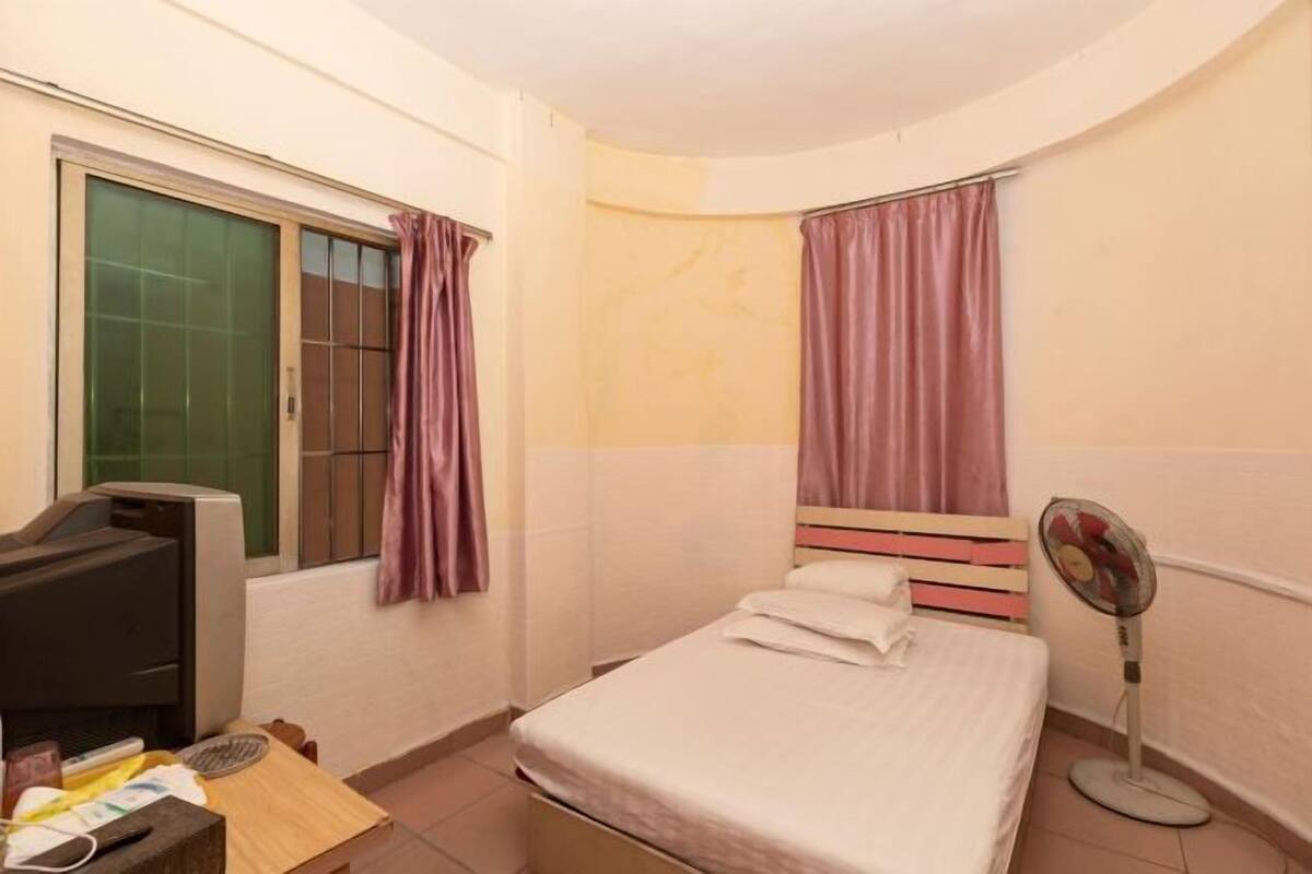 A small, single-bed room with a desk, a chair, a television, and a standing fan.
