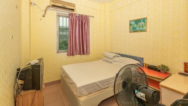 A small, neatly arranged bedroom with a bed, a desk, a chair, an air conditioner, a window with curtains, and a wall-mounted picture.