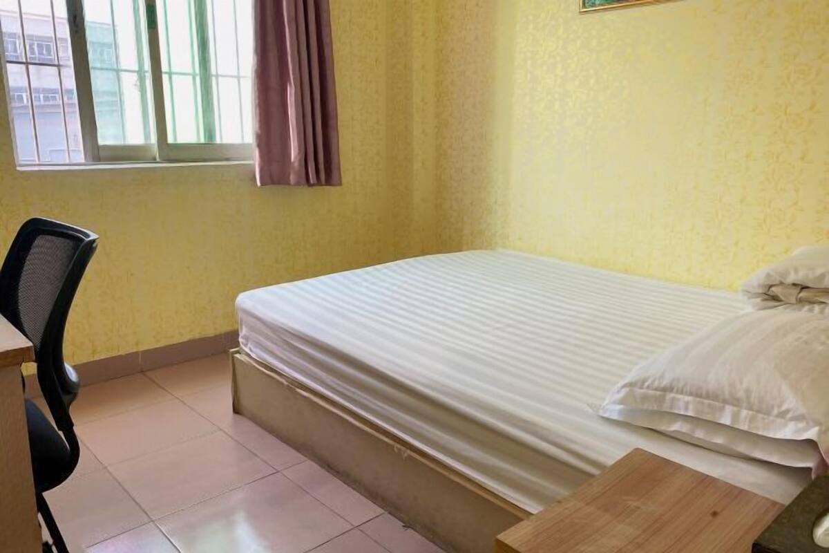 A small, single-bed room with a plain yellow wall, a window with a curtain, a desk with a chair, and a wooden nightstand.