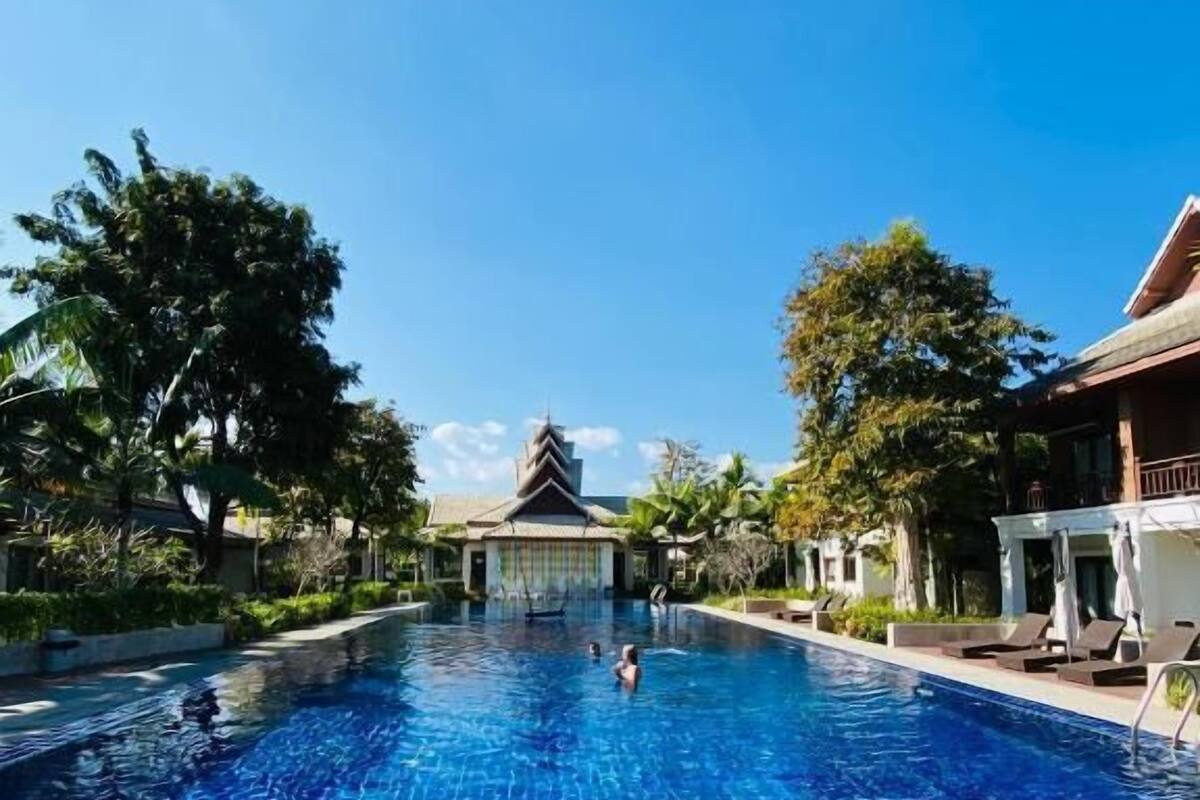 A swimming pool with tiled flooring, a person swimming, and a building with a traditional roof in the background.