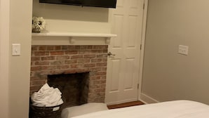 A bedroom with a brick fireplace, a mounted TV, and a white bed. - Charming Game Day House  (Opelika)
