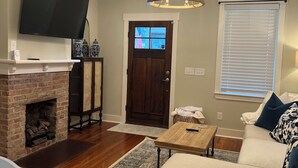 A living room with a fireplace, a television mounted on the wall, a chandelier, a sofa, and a window with blinds. - Charming Game Day House  (Opelika)