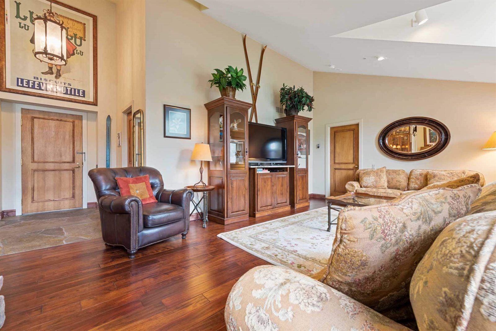 A living room with a leather armchair, a floral sofa, a wooden cabinet with a television, and a mirror on the wall.