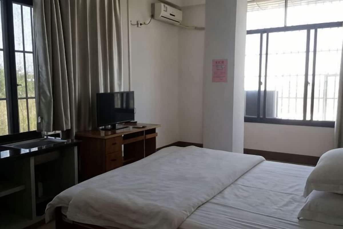 Una habitación de hotel con cama, televisor sobre un soporte de madera, ventana con cortinas y un equipo de aire acondicionado.