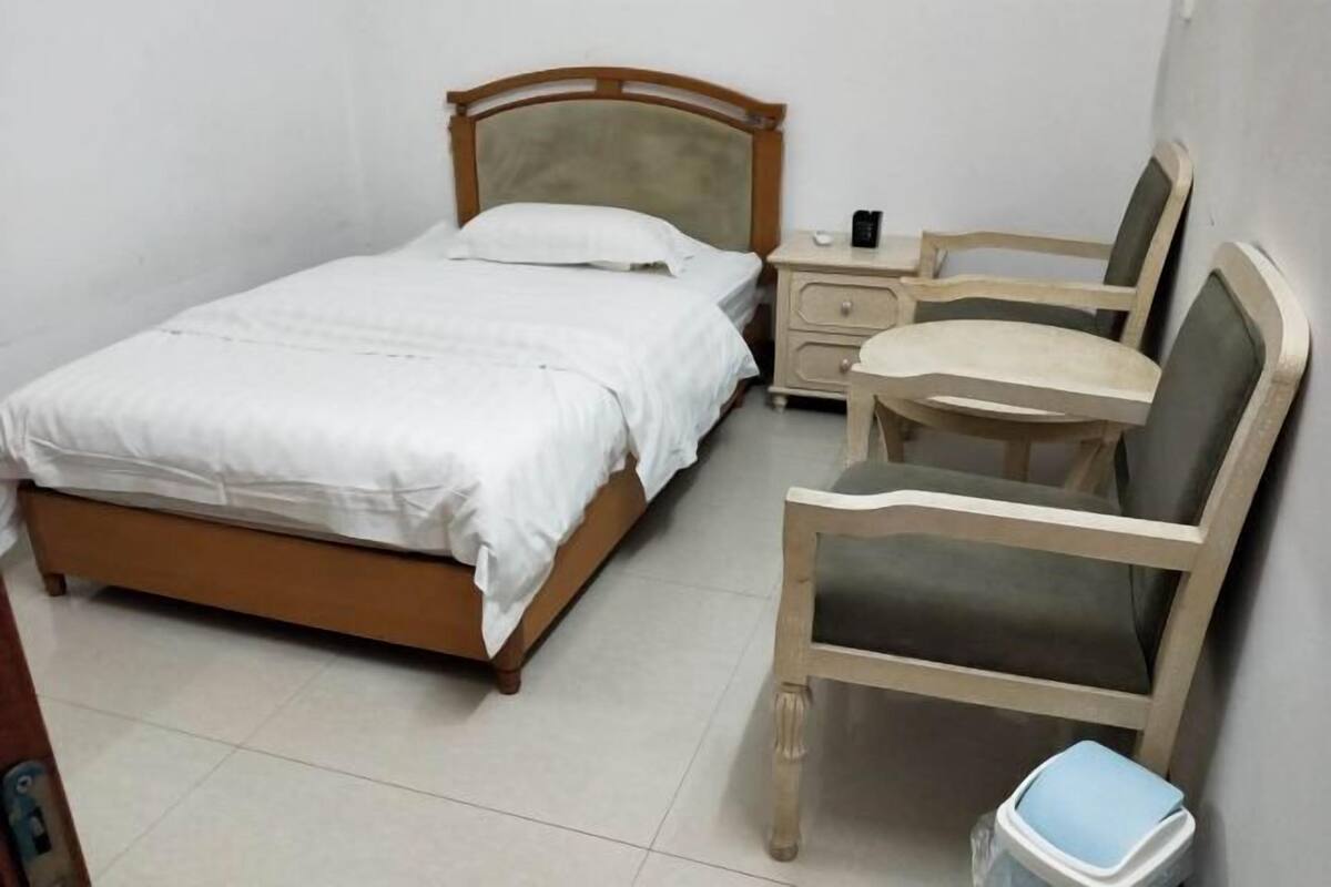 Un dormitorio con una cama individual, una mesita de noche, una silla y un cubo de basura pequeño.