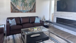 Ein modernes Wohnzimmer mit einem Ledersofa, einem Glastisch, einem Kamin und einem großen Gemälde von Palmen.