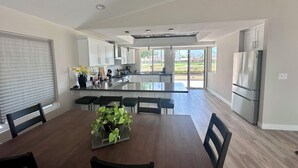 Eine moderne Küche mit einer zentralen Kochinsel, einem Essbereich mit Holztisch und -stühlen sowie Blick auf eine Wohngegend durch große Fenster.