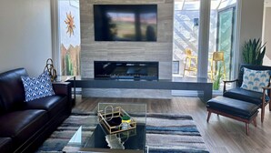 Ein modernes Wohnzimmer mit Kamin aus Stein, einem flachbildfernseher darüber, einem schwarzen Ledersofa, einem gemusterten Sessel und einem Glastisch mit Dekorationen.