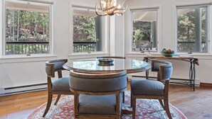 A dining area with a round glass table, four upholstered chairs, a chandelier, and large windows with a view of trees. - Beaver Creek Village 2 Bedroom Residence With Ski In/out Access and Pool (Avon)