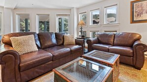 A spacious living room with a large brown leather sectional sofa, a glass coffee table, and multiple windows offering views of the outdoors. - Beaver Creek Village 2 Bedroom Residence With Ski In/out Access and Pool (Avon)