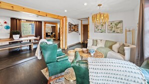 Un vaste espace de vie comprenant un canapé vert, un divan blanc et un banc en bois. Un lustre est suspendu au plafond et une grande fenêtre est agrémentée de rideaux.