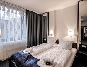 A hotel room with a bed, two bedside lamps, a mirror, and a window with curtains. - Hotel Mirage Düsseldorf-Neuss (Neuss)