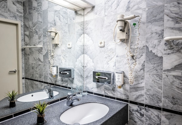 Casa de banho com azulejos de mármore, duas pias, um espelho, secador de cabelo e dispensador de toalhas.