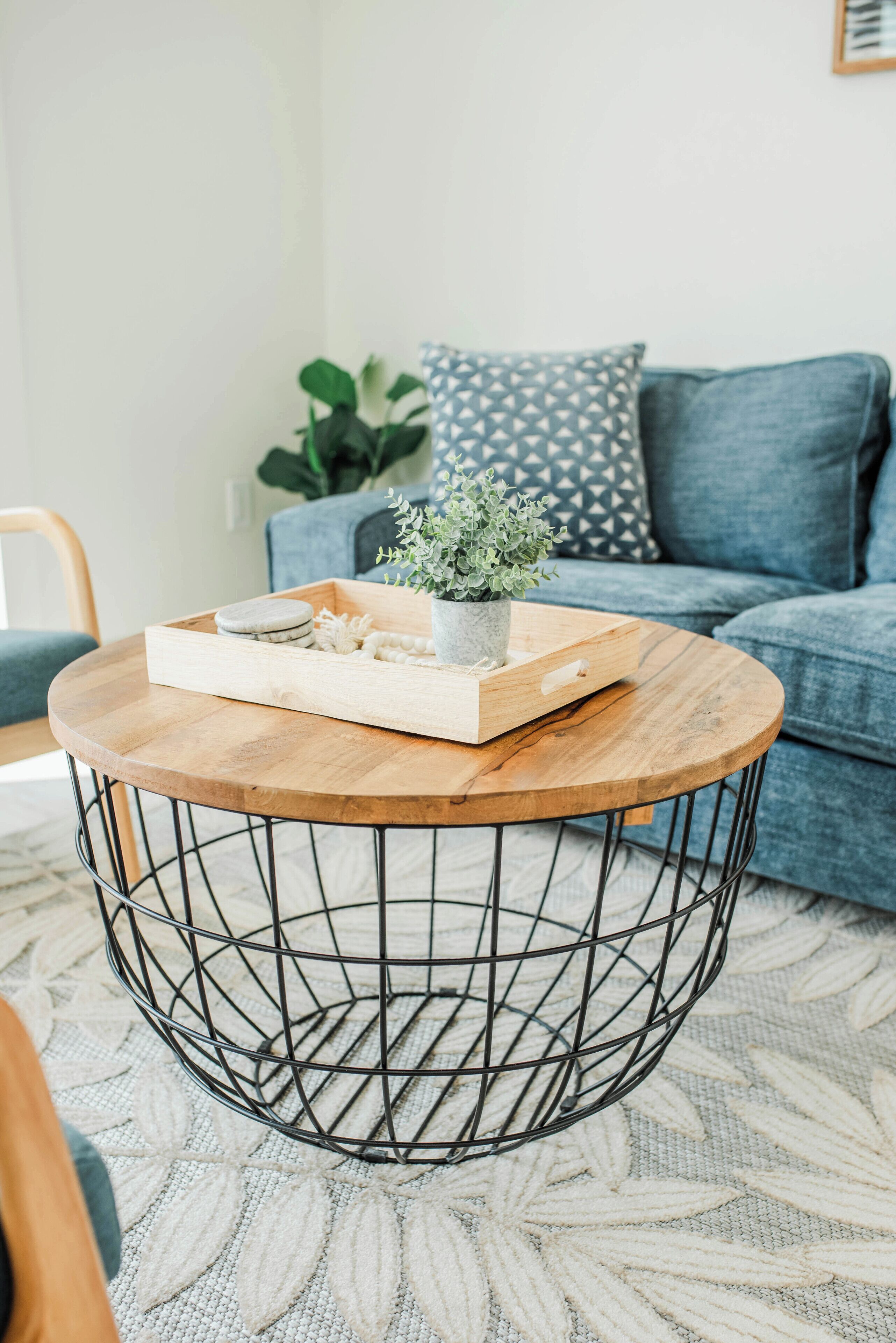 Une table basse ronde en bois, avec un pied en fil métallique, un pot de fleurs et un plateau contenant des biscuits et une bougie.