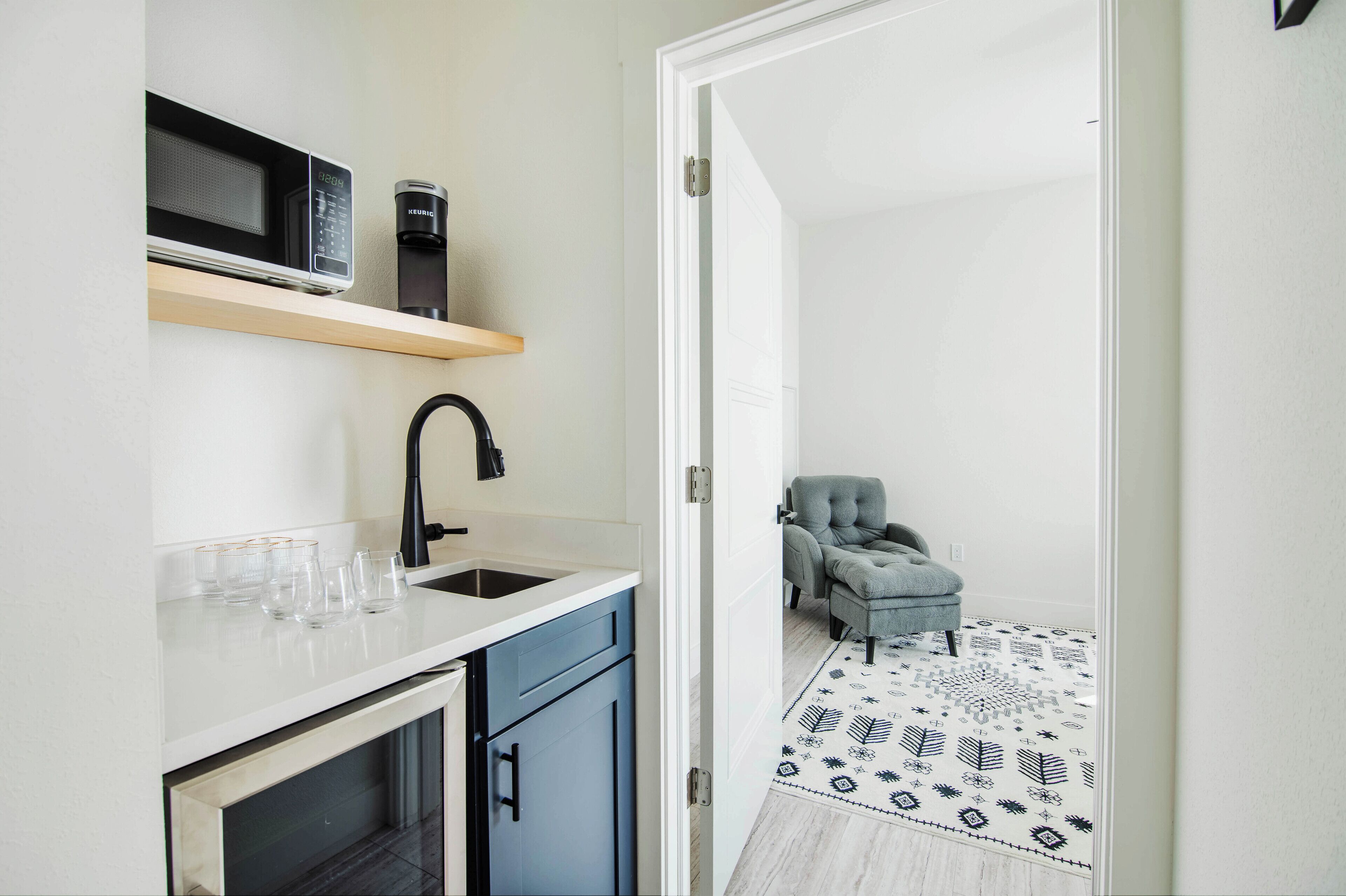 A compact kitchen with a microwave, coffee maker, and a sink. A chair is visible in the adjacent room.