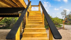Eine Holztreppe führt zu einem überdachten Bereich mit freiem Blick auf den Himmel und umliegendes Grün.