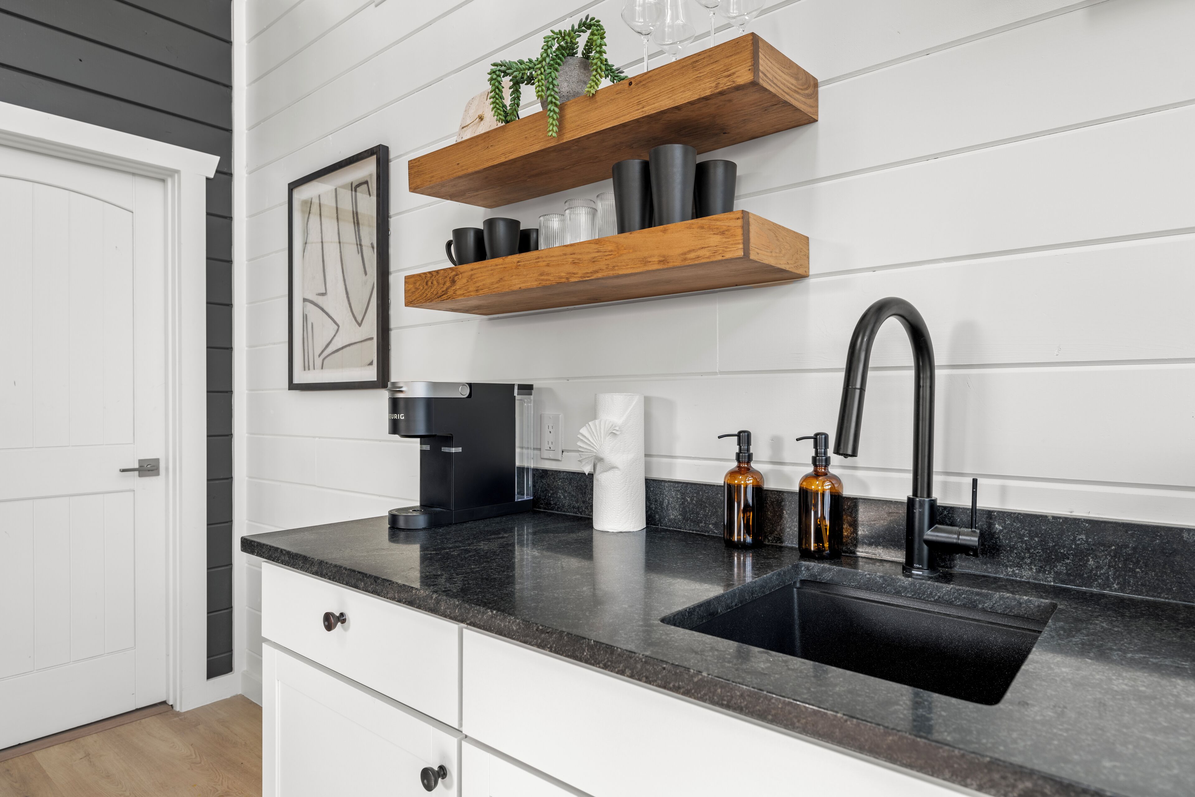 A kitchen with a black countertop, white cabinets, and a black sink. There are wooden shelves above the countertop holding various items.